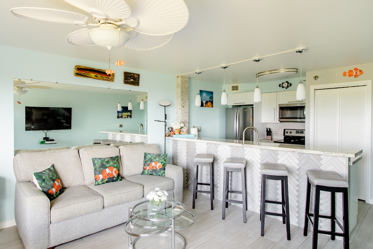 A modern living area features a light-colored sofa adorned with ocean-themed cushions. A sleek kitchen with stainless steel appliances and a breakfast bar is visible in the background, complemented by hanging pendant lights. Bright walls add to the inviting atmosphere.