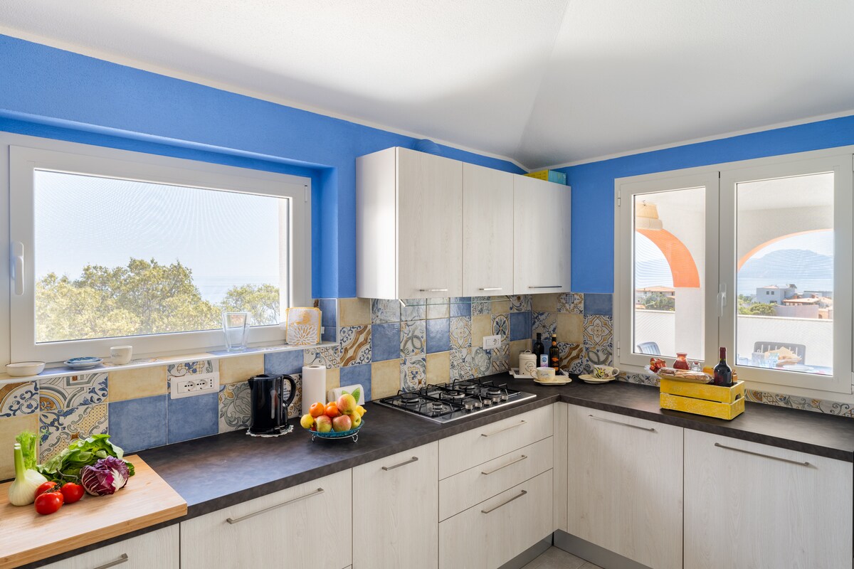 The bright kitchen features light cabinetry and a dark countertop. Two windows offer views of the sea and surrounding area. Various kitchen appliances, including a stovetop and coffee maker, are visible, along with a cutting board displaying fresh fruits and vegetables.
