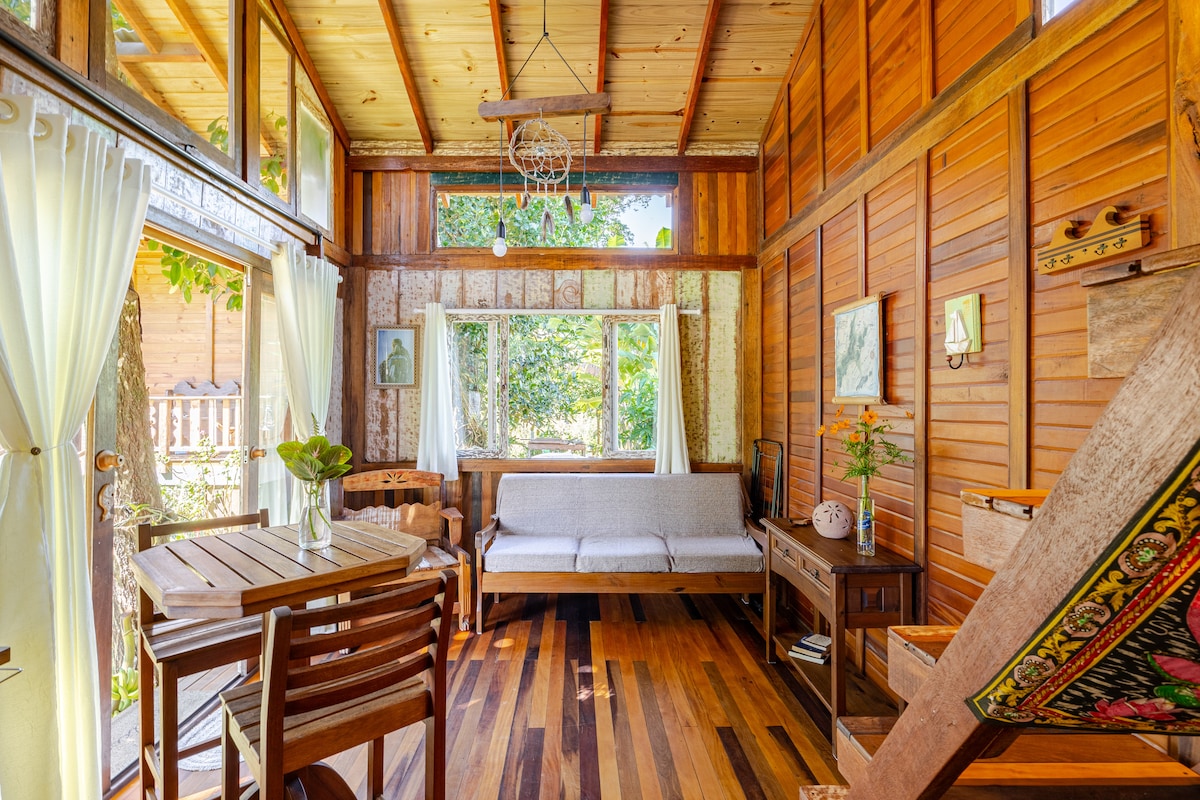 A cozy living area features wooden walls and floors, complemented by large windows allowing ample natural light. A simple sofa sits alongside a dining table made of wood. Decorative elements, including plants and artwork, add a touch of tranquility to the space.