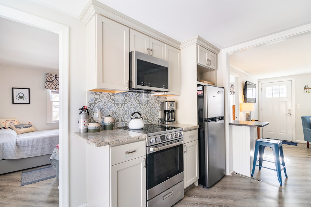 A modern kitchen area is featured, equipped with stainless steel appliances, including a microwave and refrigerator. The cabinetry is a soft neutral tone, complemented by decorative tile backsplash. An island bar with tall blue stools provides seating, while natural light enters from adjacent spaces.