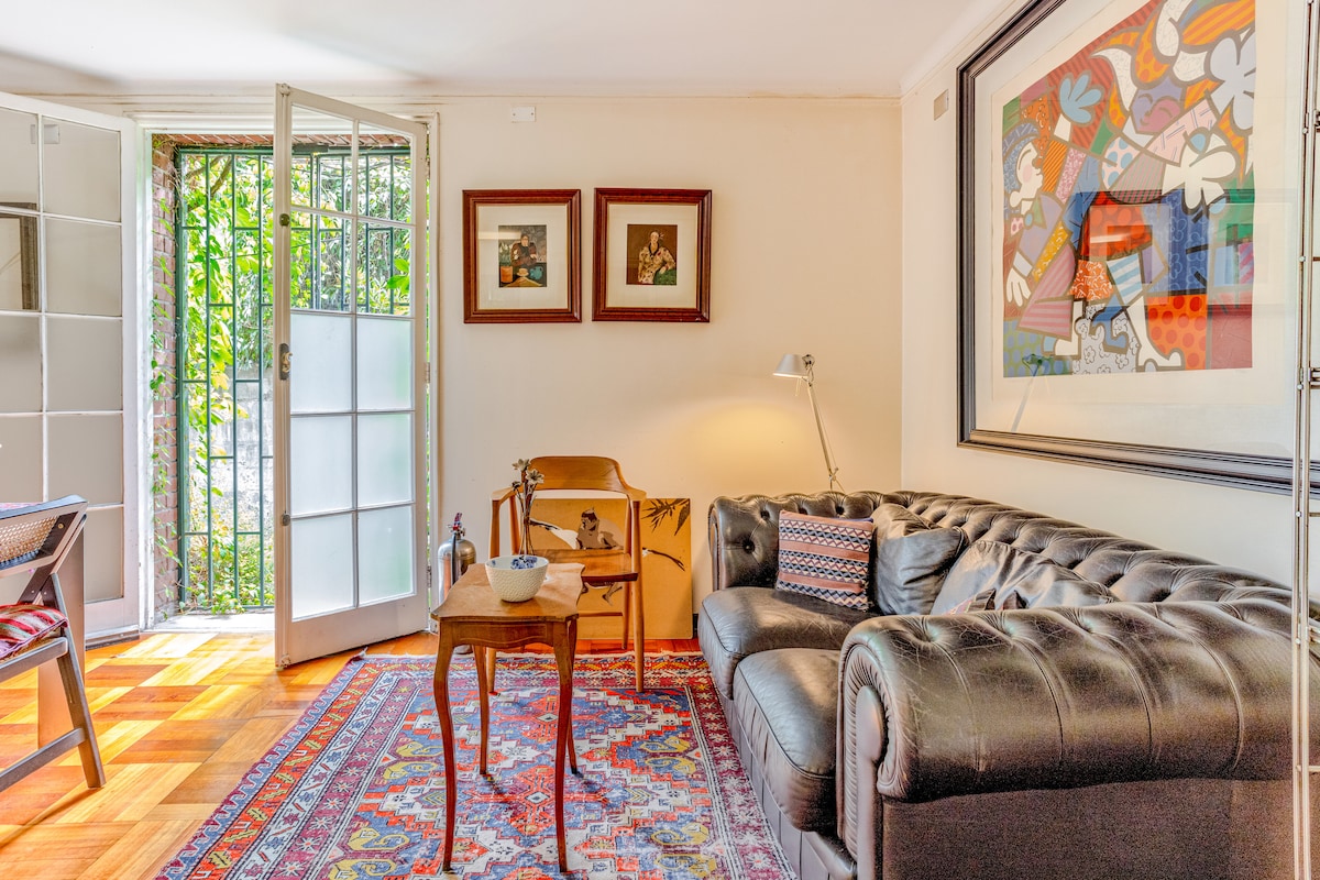 A cozy living area is highlighted by a dark leather sofa and a vibrant area rug. Natural light filters in through open glass doors that lead to a green outdoor space. Wall art and framed pictures add character to the warm ambiance.