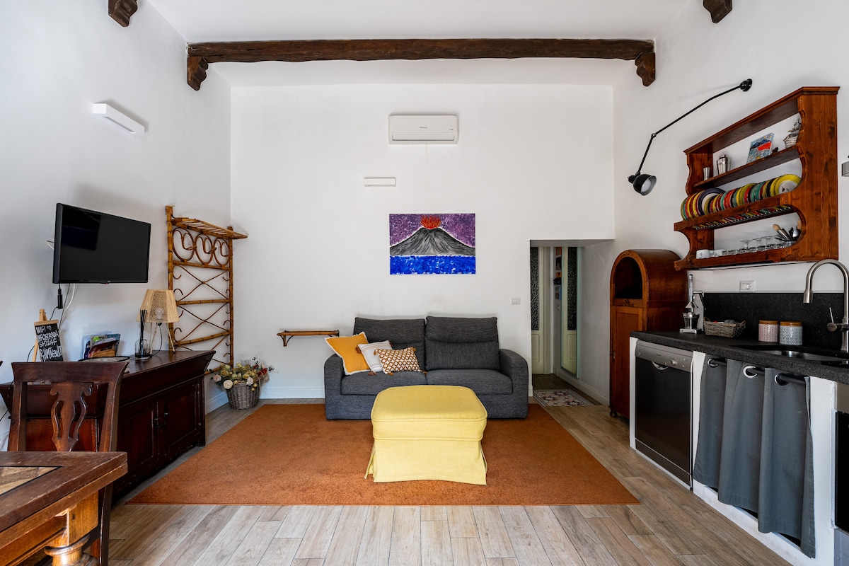 The living area is spacious and well-lit, featuring a comfortable gray sofa and a vibrant yellow throw blanket. An artistic mountain painting adorns the wall, while a wooden shelf displays colorful dishware. A kitchenette is visible in the background, highlighting convenient amenities.
