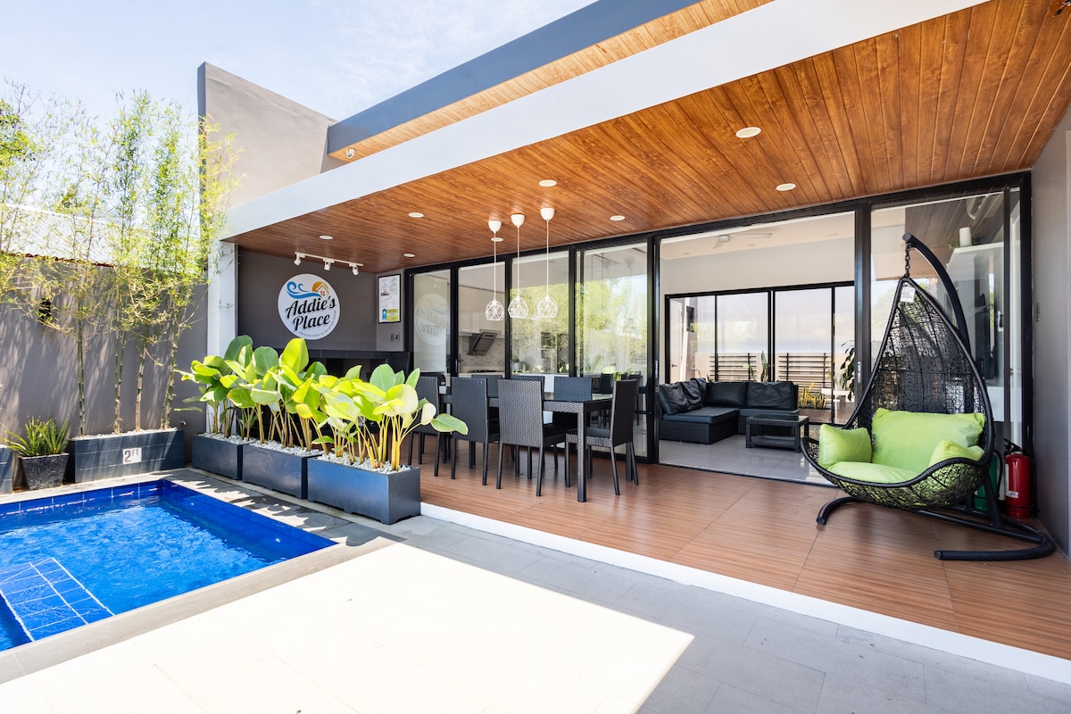 An outdoor living space features a modern design with sliding glass doors that open to a private pool. A dining area accommodates multiple guests, while lush plants add greenery. A hanging chair provides a comfortable seating option, enhancing the inviting atmosphere of the space.