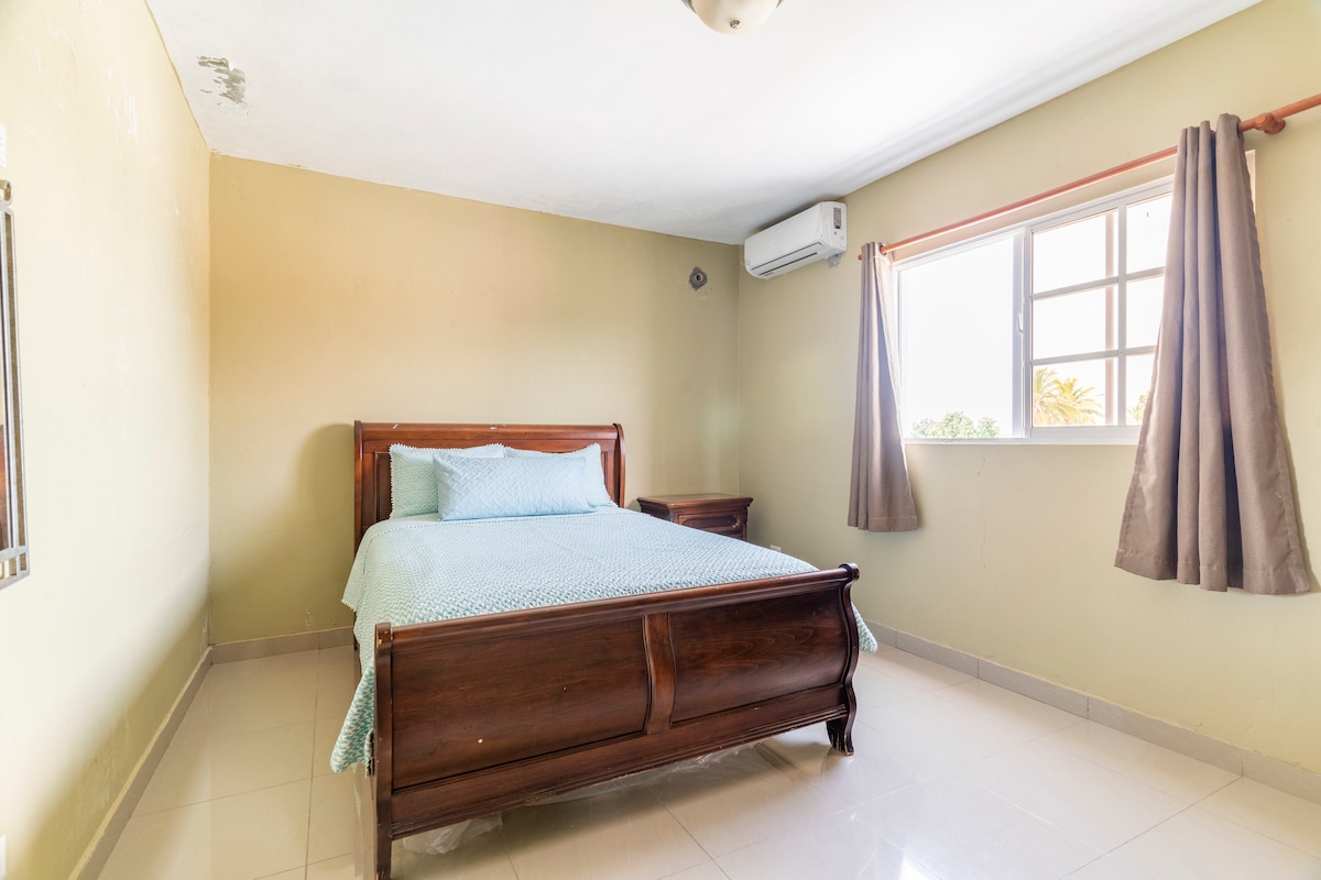 A bedroom features a wooden bed with a light blue coverlet. A nightstand is positioned beside the bed. Natural light enters through a window, which is dressed with light curtains. An air conditioning unit is mounted on the wall near the ceiling.