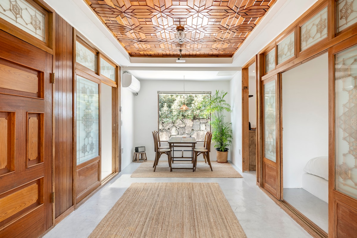 A spacious interior features wooden doors and an intricately designed ceiling with geometric patterns. A dining table with chairs is positioned in a bright, airy space, surrounded by plants and natural light filtering through large windows. The floors are covered with soft, neutral-toned textures.
