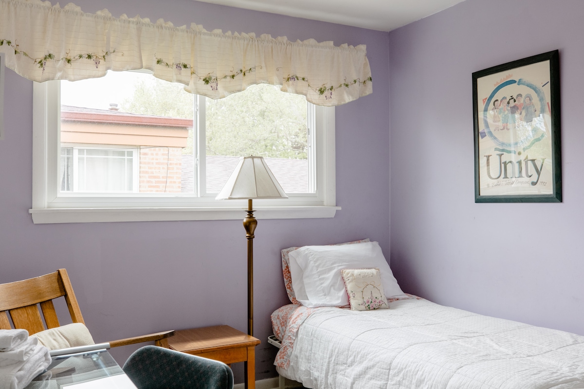 A purple-walled bedroom is furnished with a spacious bed layered with white bedding. A small table is positioned beside the bed, with a lamp illuminating the space. A window is adorned with a floral curtain, allowing natural light to brighten the room.