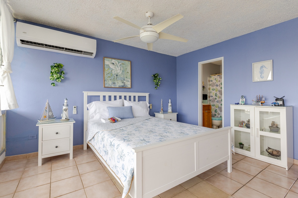The bedroom features a spacious bed adorned with light blue bedding, complemented by a decorative white headboard. A fan is mounted on the ceiling, providing comfort. Two nightstands flank the bed, while a glass cabinet displays decorative items. Natural light enters through the window, enhancing the serene atmosphere.