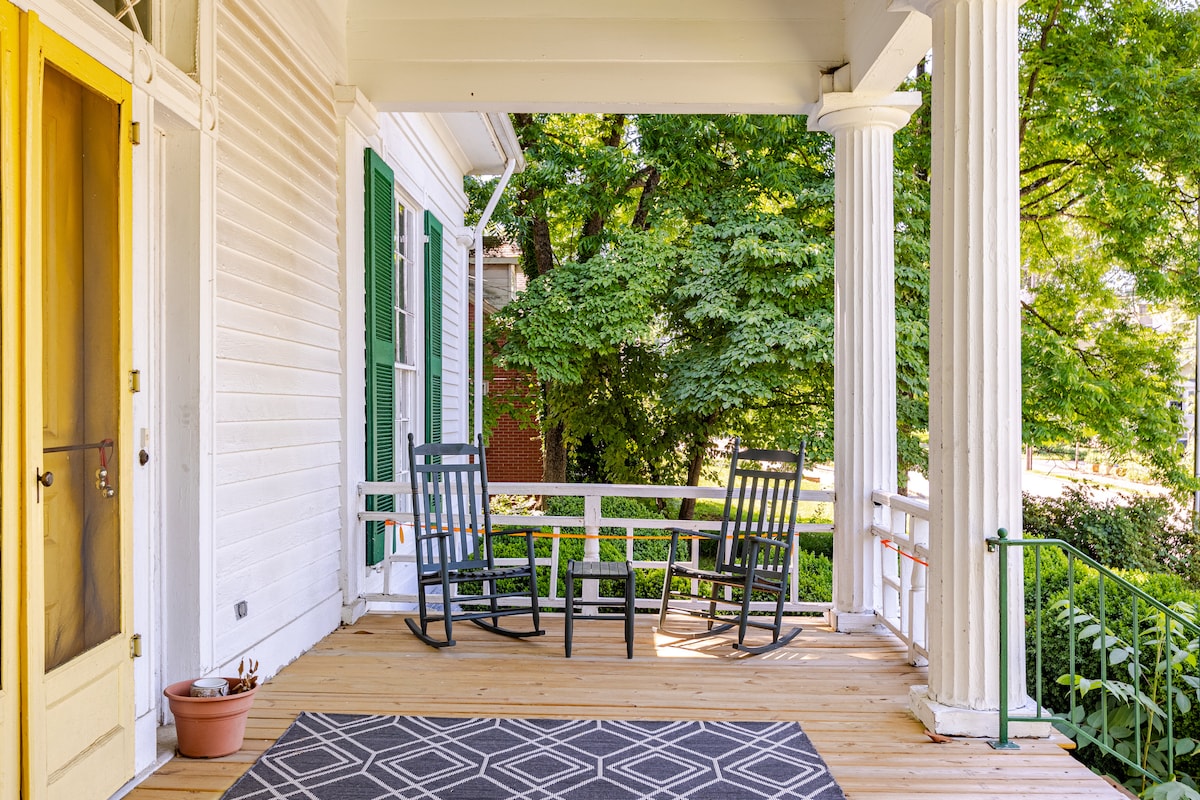 A spacious front porch features wooden flooring and a patterned area rug placed in the center. Two rocking chairs and a small table are positioned for relaxing. Lush greenery from surrounding trees enhances the tranquil atmosphere.