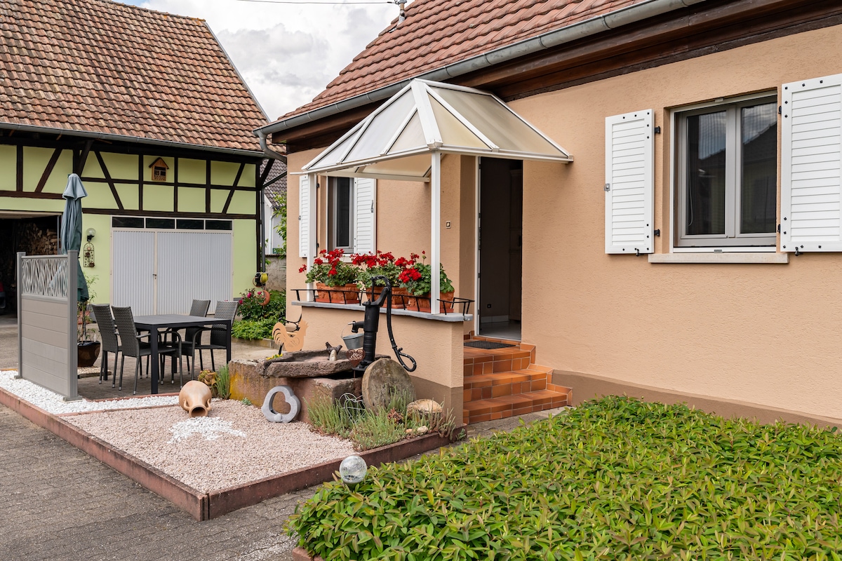 The exterior of the house features a welcoming entrance with a covered porch and potted red flowers. A wooden table and chairs are arranged on the patio, accompanied by a large umbrella. Fresh greenery surrounds the area, enhancing the inviting aspect of the home.