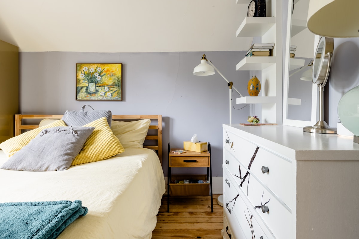 A cozy bedroom features a wooden bed adorned with several textured pillows. A small bedside table holds a reading lamp and a decorative item. A white dresser is positioned against the wall, while a painting adds a splash of color to the room.