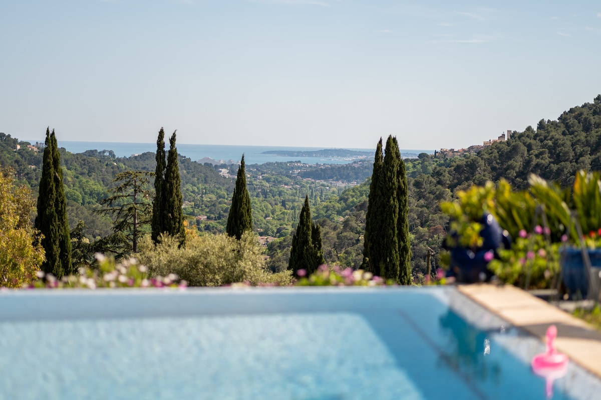 A scenic view reveals a swimming pool in the foreground, bordered by vibrant greenery and colorful flowers. Towering cypress trees frame the vista, which extends to rolling hills and the distant coastline, showcasing the tranquility of the surrounding landscape.