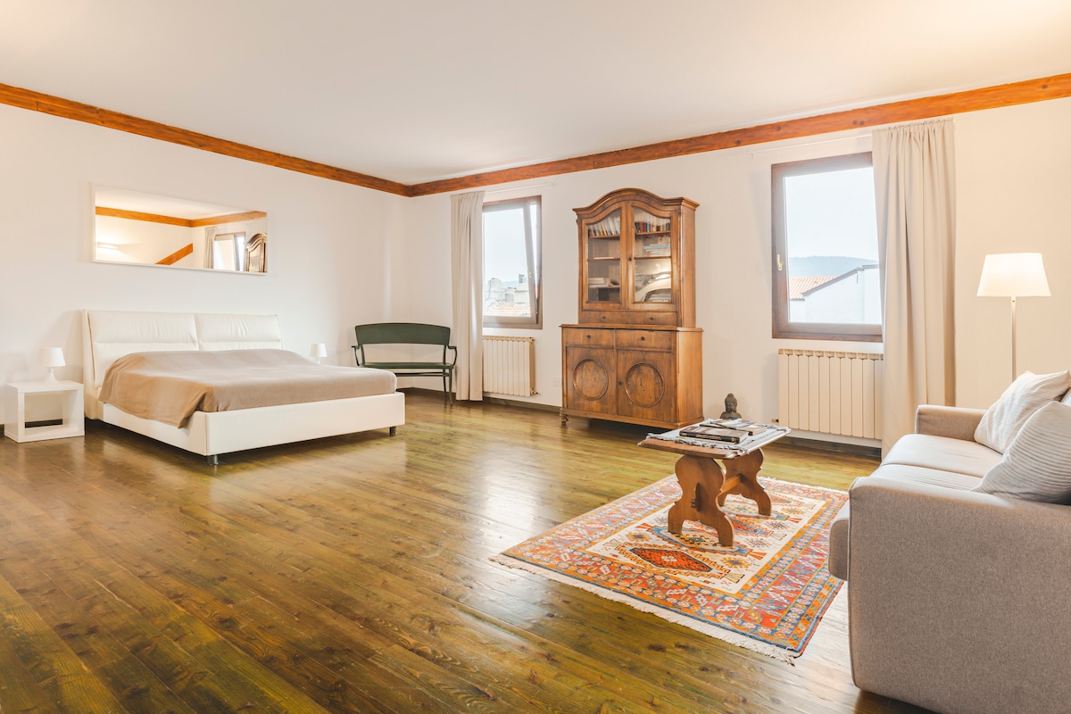 A spacious room features a comfortable bed with neutral bedding. A vintage wooden cabinet stands against the wall, complemented by a soft sofa and an intricately designed area rug. Large windows allow natural light to fill the space, enhancing the warm wood flooring.