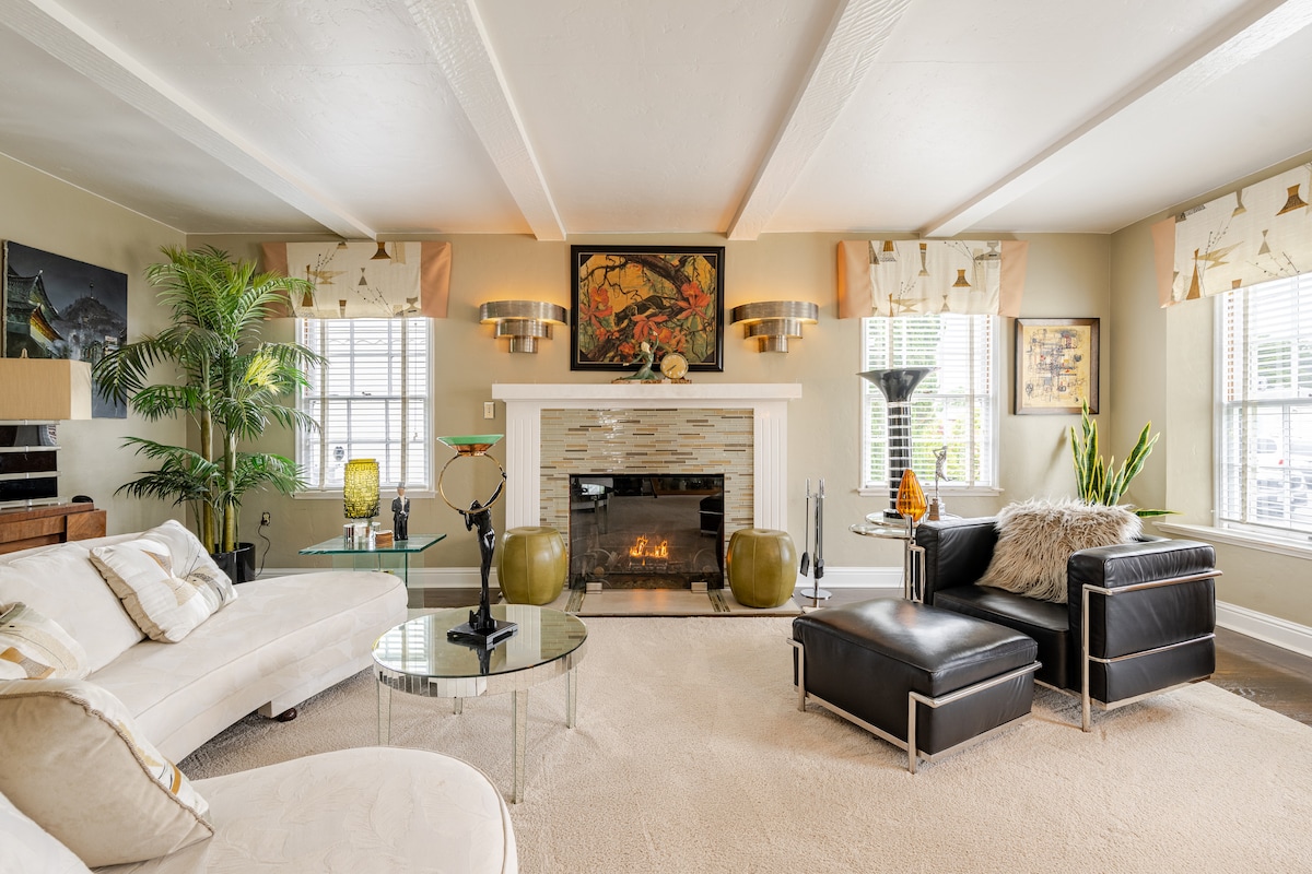 A comfortable living area features a white sectional sofa and a sleek black lounge chair paired with a glass coffee table. Natural light pours in through large windows adorned with soft drapes. A stone fireplace serves as a focal point, surrounded by decorative artwork and plants.