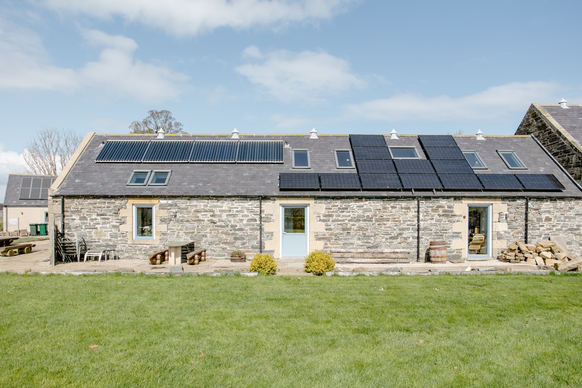 The exterior of the steading conversion showcases a traditional stone structure, enhanced with solar panels on the roof. Lush green grass surrounds the building, complemented by wooden benches and planters. Large windows provide ample natural light, reflecting the serene rural setting.