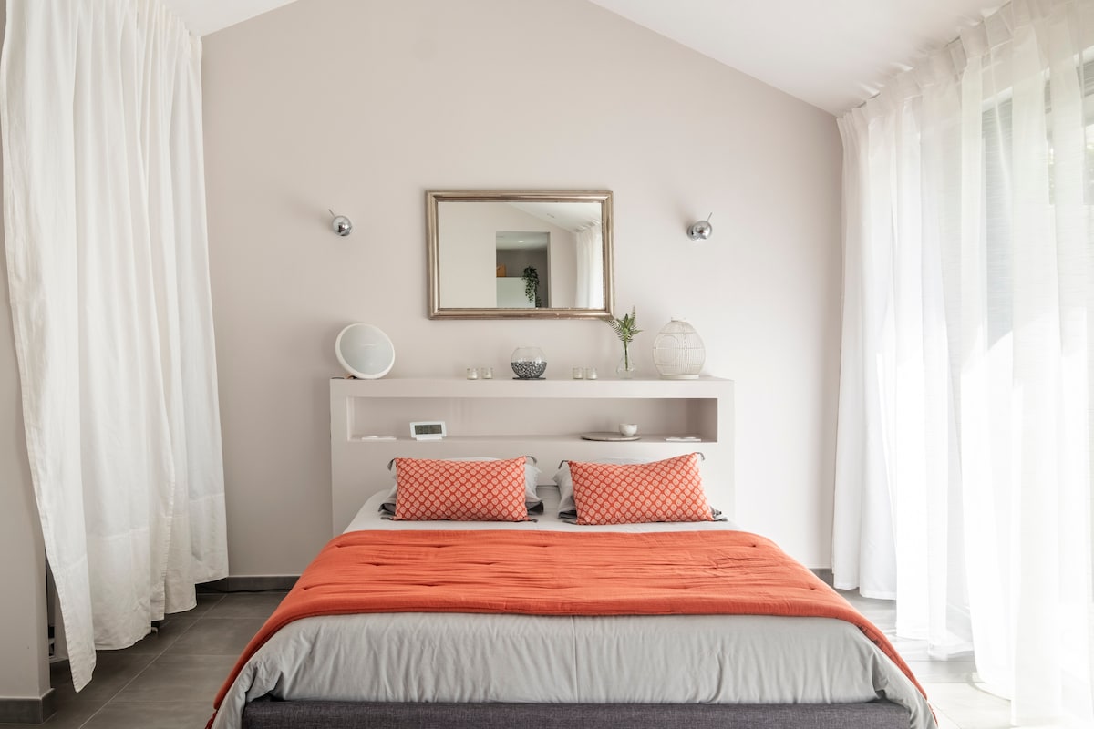 A well-appointed sleeping area features a spacious bed adorned with an orange throw and decorative pillows. A minimalist headboard is complemented by a large mirror above it, reflecting natural light from the adjacent window, which is dressed with sheer white curtains.