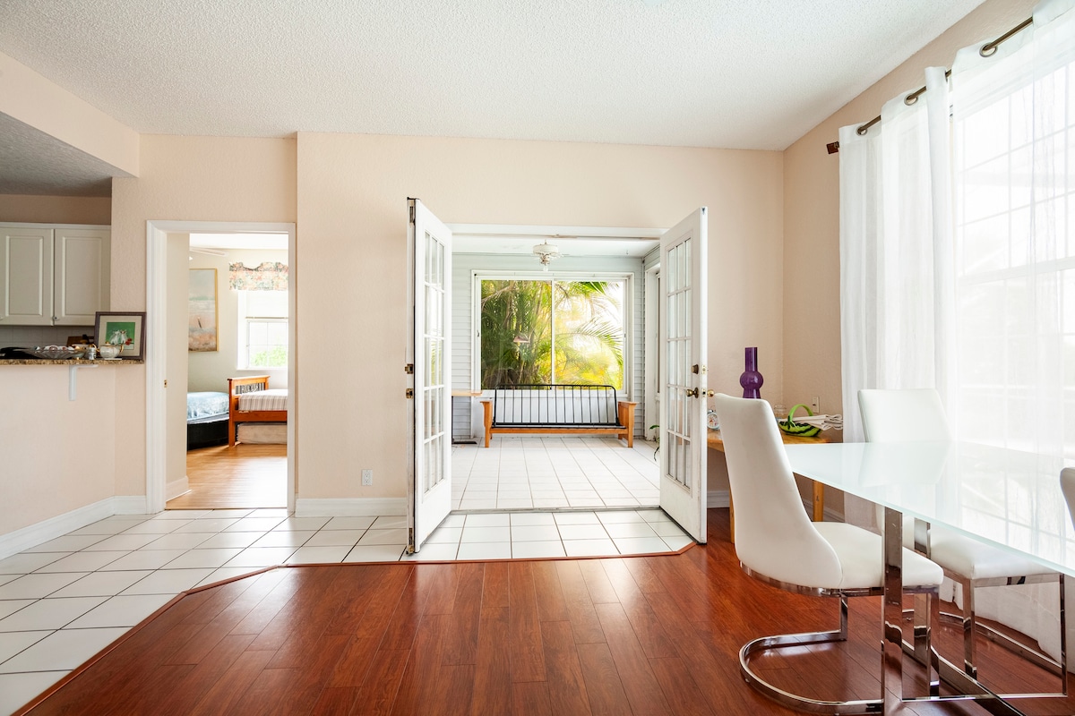 An open layout is presented, connecting a bright dining area with a modern table and white chairs to a sunlit living space. French doors frame a view of a private sunroom adorned with tropical plants, enhancing the sense of spaciousness and tranquility.