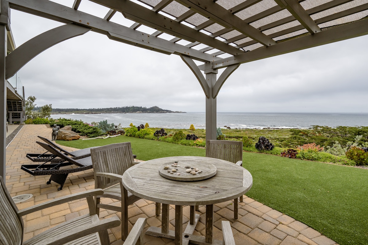 An outdoor patio area is presented, featuring a wooden dining table surrounded by several chairs. The view reveals the ocean and coastline under a soft, cloudy sky, with a well-maintained lawn and landscaped garden visible in the foreground.