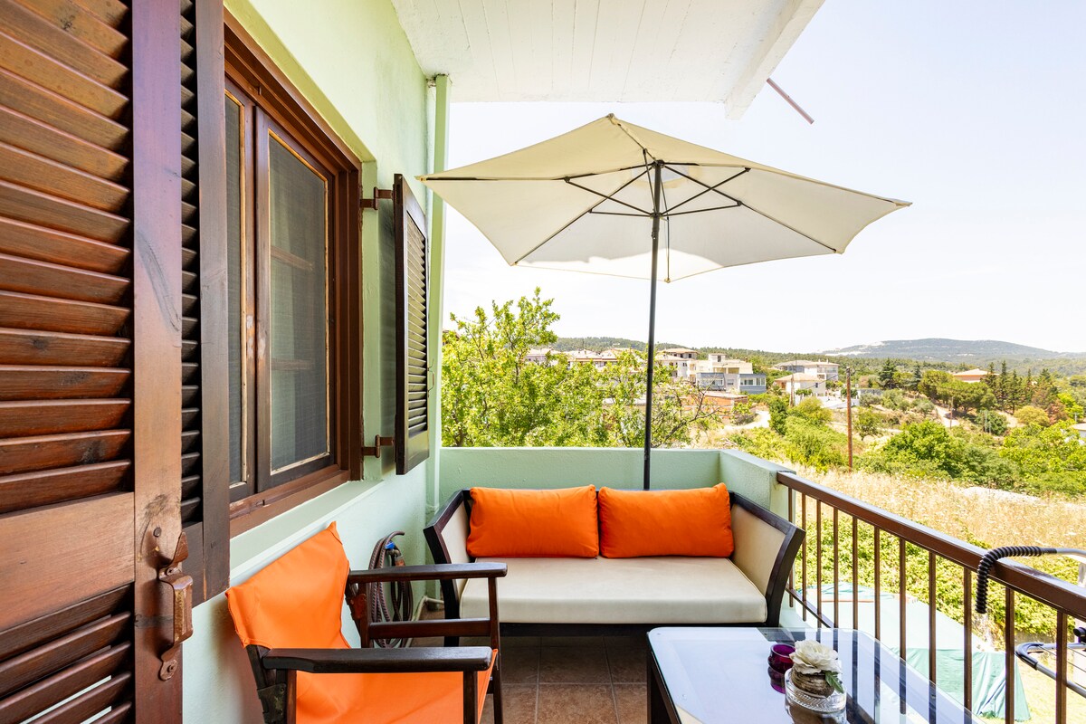 A balcony equipped with a comfortable seating arrangement features a sofa adorned with orange cushions alongside two wooden chairs. A large umbrella provides shade, creating a serene outdoor space. Lush greenery and distant hills are visible beyond the railing.