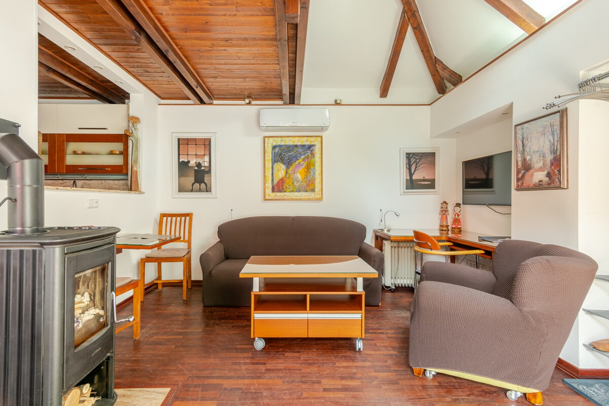 A comfortable living area features wooden beams and a mix of modern and classic furniture. A large sofa and armchair are positioned around a wooden coffee table. Artworks adorn the walls, and a small desk is situated in the corner, offering a functional workspace.