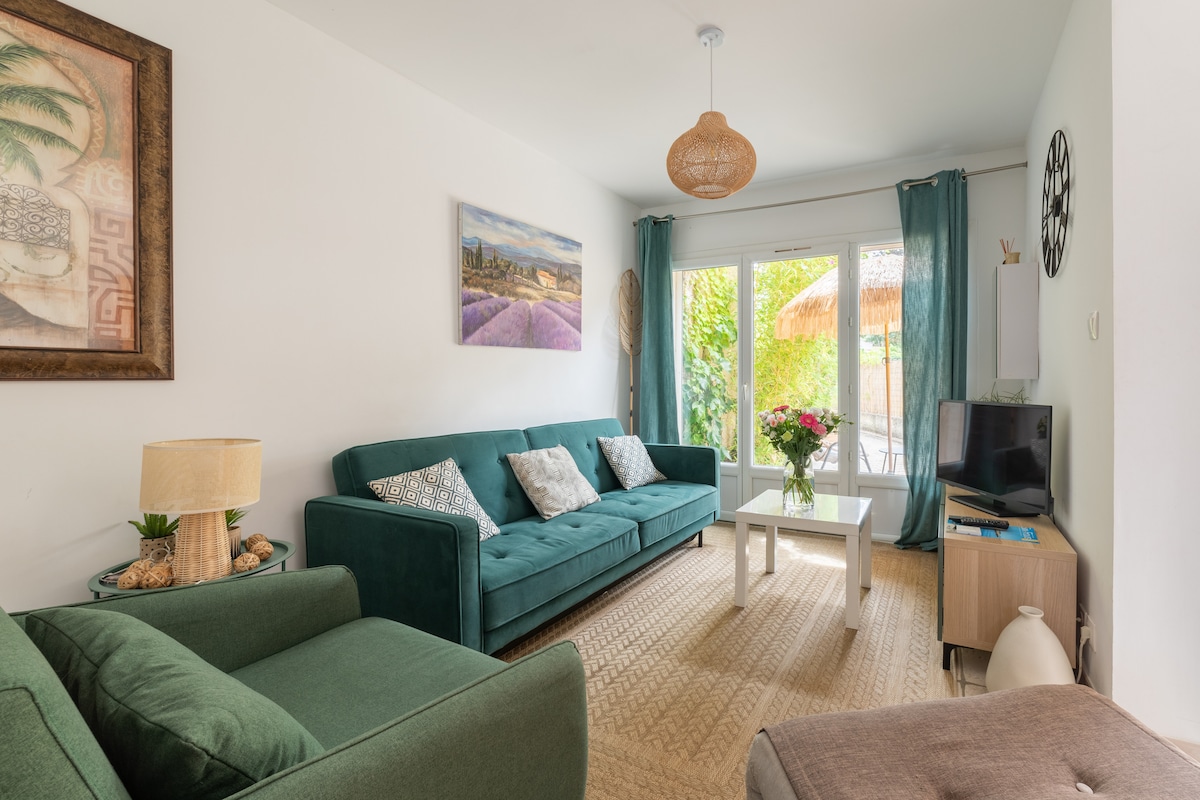 A living area features a green sofa and armchair, complemented by a white coffee table. Natural light filters through large glass doors, opening to a garden space. A decorative artwork adorns the wall, while a small flower arrangement adds a touch of nature.