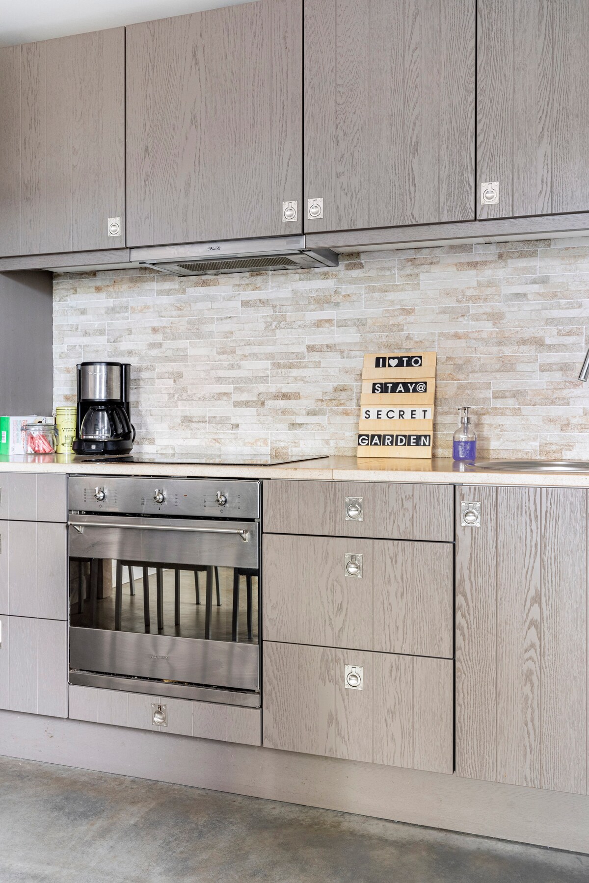 An organized kitchen features modern cabinets in light wood with sleek handles. A stainless steel oven is set beneath a countertop, with a coffee maker positioned nearby. The stone backsplash adds texture, while a sign reading 'NOTO STAY SECRET GARDEN' is displayed on the counter.