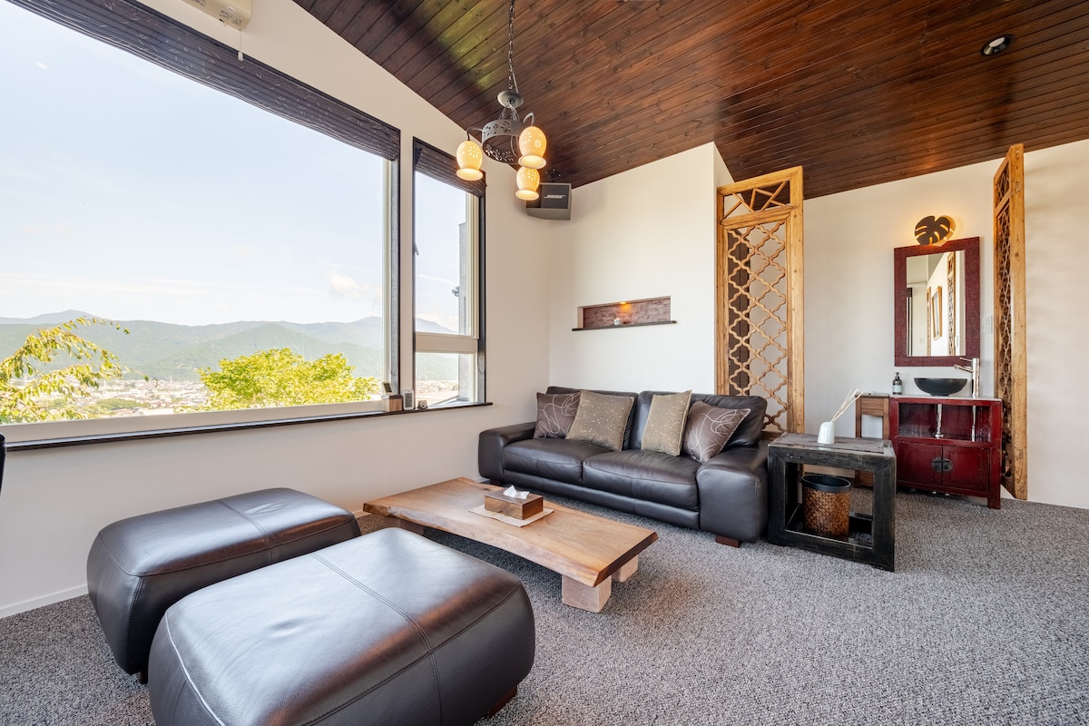 A modern living area features large windows that provide panoramic views of the surrounding mountains and city. A dark leather sofa and matching ottomans complement a rustic wooden coffee table. The walls are adorned with decorative accents, adding a touch of elegance to the space.