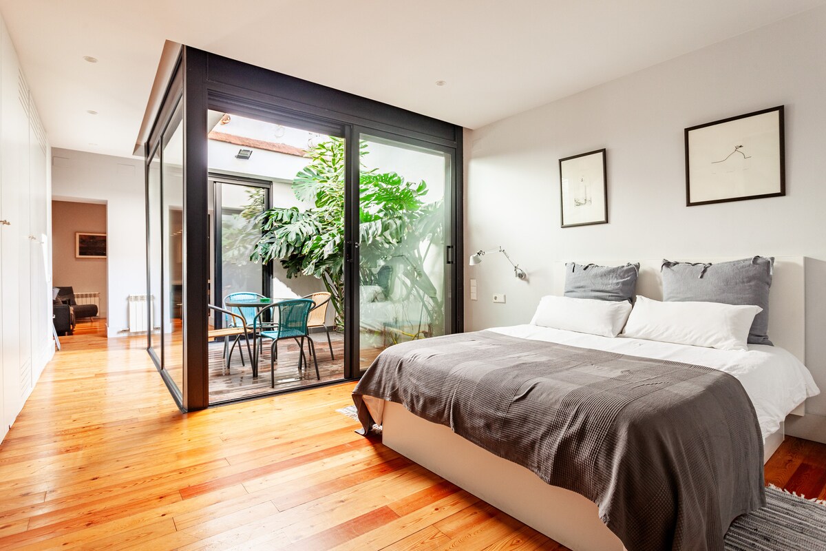 A well-lit bedroom features a spacious bed with a gray blanket, flanked by modern sconces. Floor-to-ceiling glass doors open to a small terrace with green plants and outdoor seating. The warm wooden flooring complements the calming color scheme throughout the space.