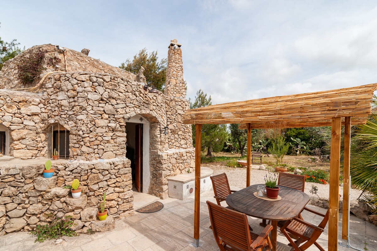 An exterior view of a rustic stone trullo is presented, featuring a small porch with a wooden pergola. A round table with four wooden chairs is set beneath the cover, surrounded by potted cacti and a garden area with various plants in the background.