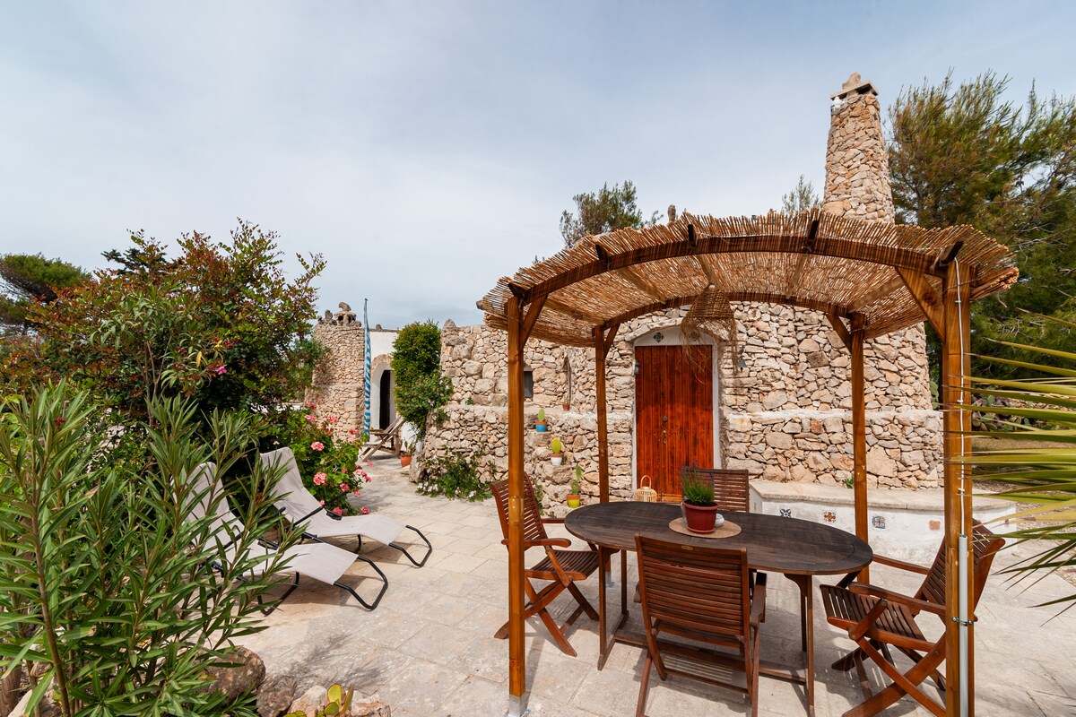 A shaded patio area features a wooden table surrounded by four chairs, with a potted plant on the table. Natural stone walls form the backdrop, and sun loungers are positioned nearby, complemented by lush greenery and flowering plants in the surrounding garden.