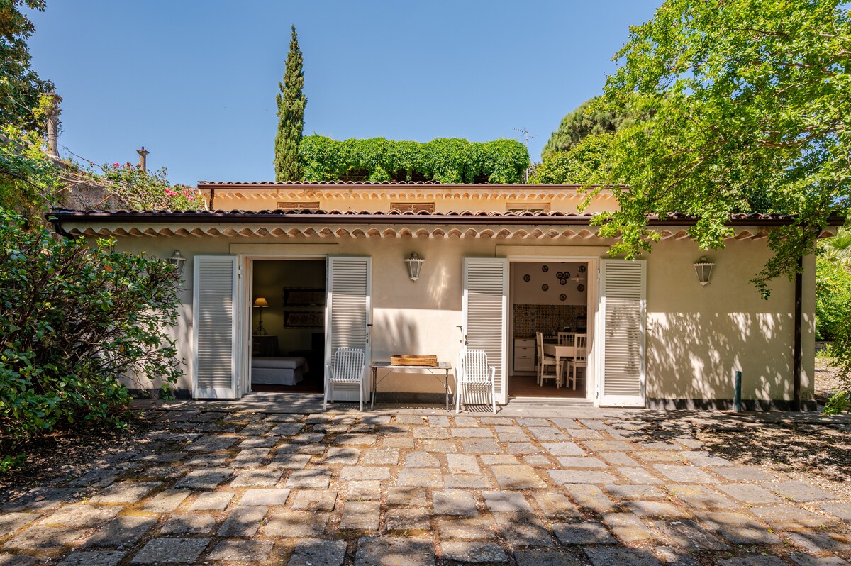 The outbuilding features a spacious patio with stone pavers, surrounded by well-kept greenery. White shutters frame the open doors, revealing an inviting interior. A wooden table and two chairs are positioned outside, providing a relaxing space to enjoy the surrounding garden.