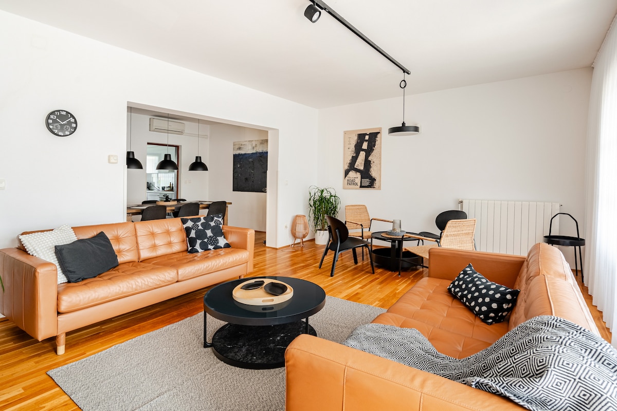 A well-lit living room features two stylish leather sofas, a round coffee table, and a dining area visible in the background. Light wooden flooring complements neutral walls. Decorative cushions add detail, while large windows ensure a bright, spacious atmosphere.