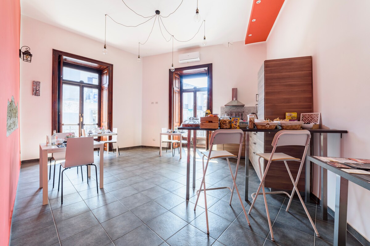 A bright dining area features several tables arranged neatly, with chairs positioned for guest comfort. Natural light fills the space through large windows. A small kitchenette is visible in the corner, equipped for meal preparation. The walls are painted in soft colors, creating a welcoming atmosphere.