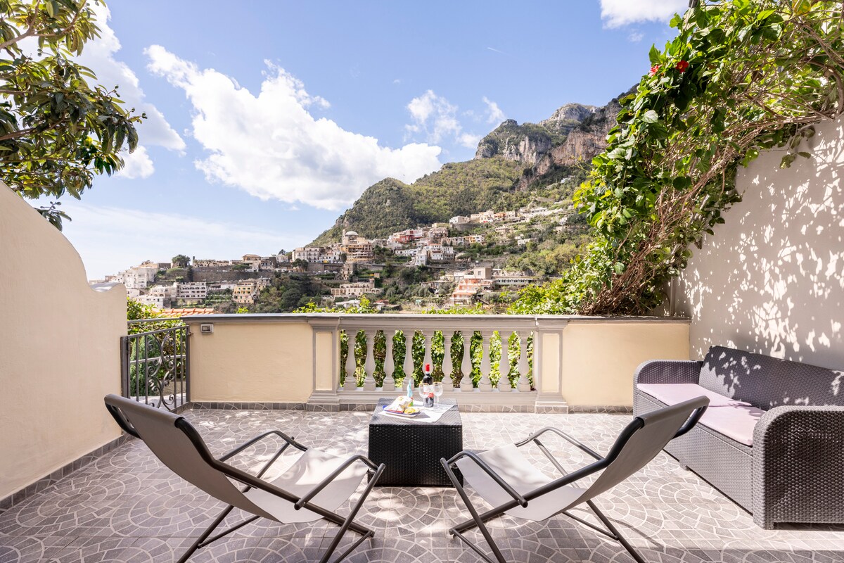 A spacious terrace offers panoramic views of the surrounding hills and city. Two lounge chairs are positioned beside a small table, creating a relaxing outdoor space. Lush greenery borders the area, enhancing the natural light from the clear blue sky.