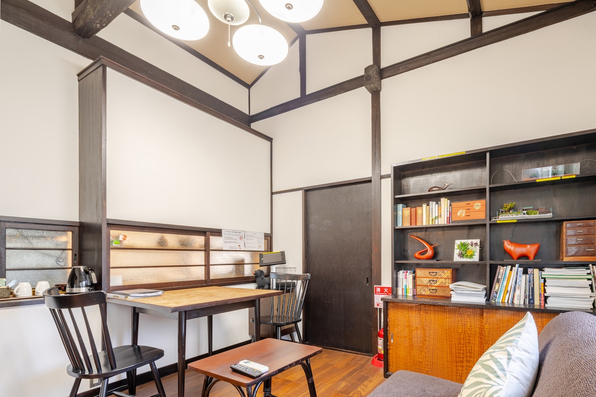 The study space features a wooden table surrounded by black chairs, complemented by an antique desk with bookshelves filled with various books. Soft lighting is provided by a modern ceiling fixture, enhancing the functionality of this tranquil workspace.