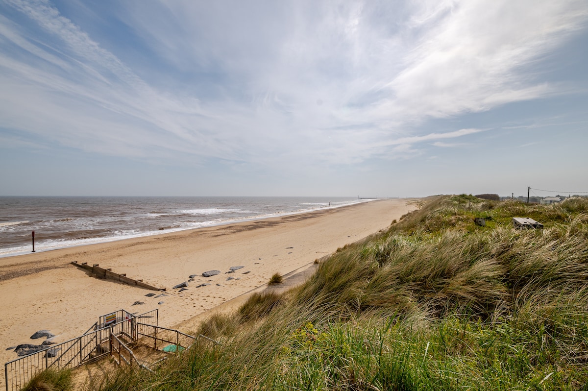 A sandy beach is visible with gentle waves lapping at the shore. Dunes with tall grass frame the edge of the beach, leading towards the horizon. A few weathered wooden posts are scattered along the shoreline, and a fence is seen at the top of the dunes.