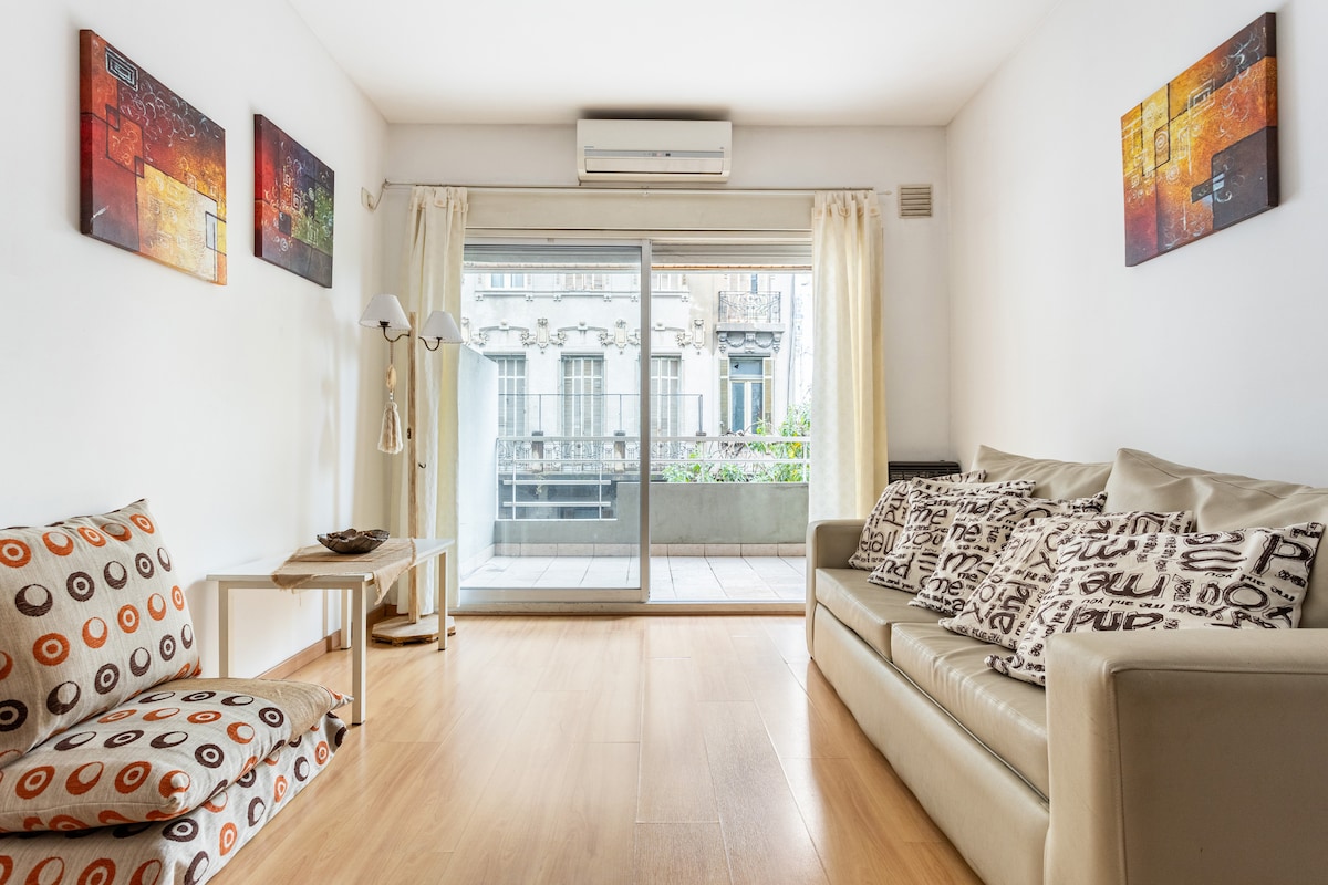 A bright living area presents a comfortable sofa adorned with patterned cushions. A secondary seating option is visible alongside a small table. Large glass doors lead to a balcony, allowing natural light to fill the room. Wall art adds subtle color to the white walls.