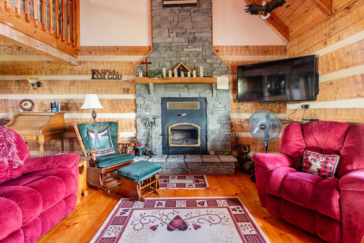 A rustic living area features a stone fireplace as the focal point, surrounded by wooden walls and a warm color palette. Two red recliners and a green rocking chair offer seating options, while a decorative area rug adds comfort underfoot. Natural light streams in through nearby windows.
