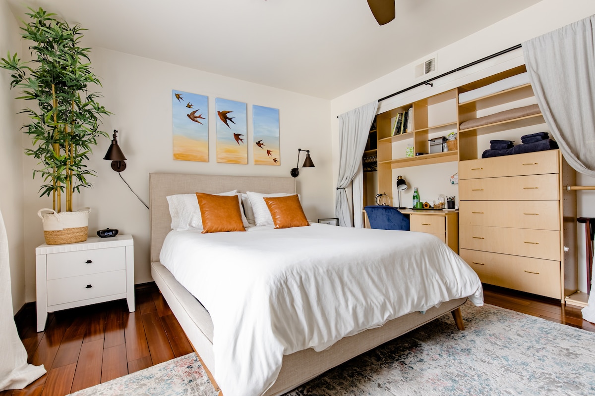 A serene bedroom features a spacious bed with white linens and decorative pillows. Two bedside lamps are positioned on either side of the bed. A vibrant wall art trio depicting birds in flight adorns the wall above the bed. Natural light is filtered through soft curtains, complementing the warm wood flooring.