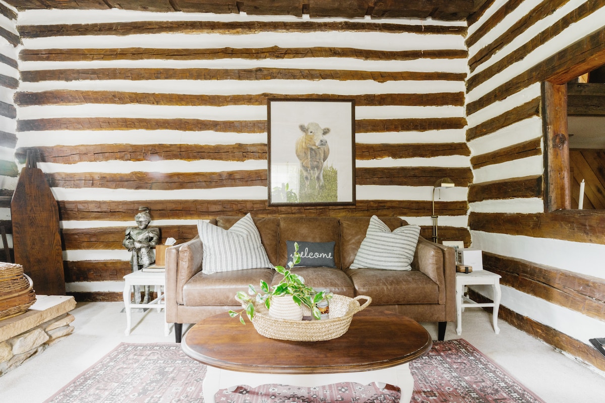 A cozy living room is framed by rustic log walls, featuring a comfortable brown sofa adorned with patterned cushions. A round coffee table with a potted plant sits in the center. A framed artwork of a cow adds character to the space.