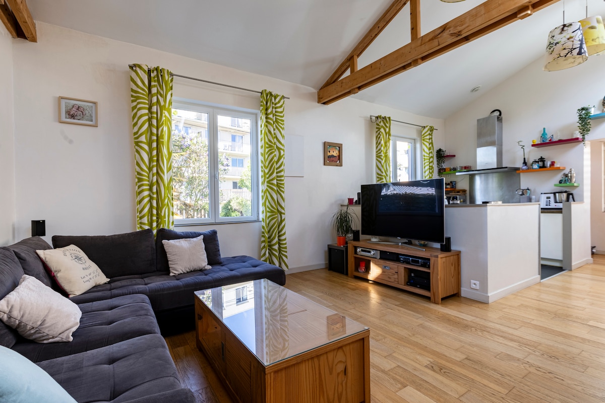 A bright living area features a dark sectional sofa with decorative cushions, a glass-top coffee table, and a large television on a wooden stand. Sunlight streams through windows dressed with leafy green curtains, illuminating the space. An open kitchen in the background is equipped with modern appliances.