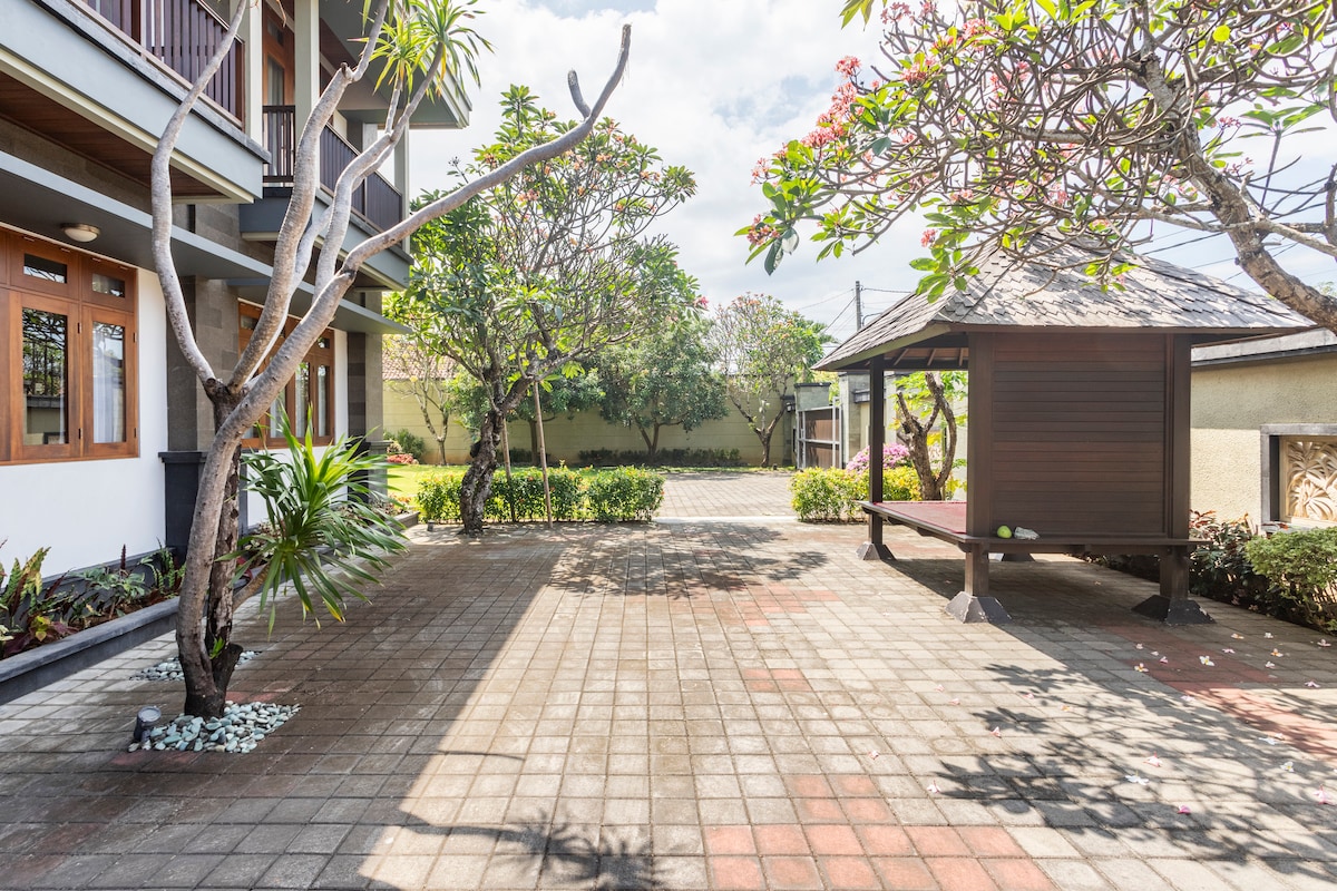An outdoor area features a paved surface surrounded by lush greenery and blooming trees. A traditional shaded gazebo is visible, providing a space for relaxation. The environment is calm, complemented by gentle natural light that filters through the leaves.
