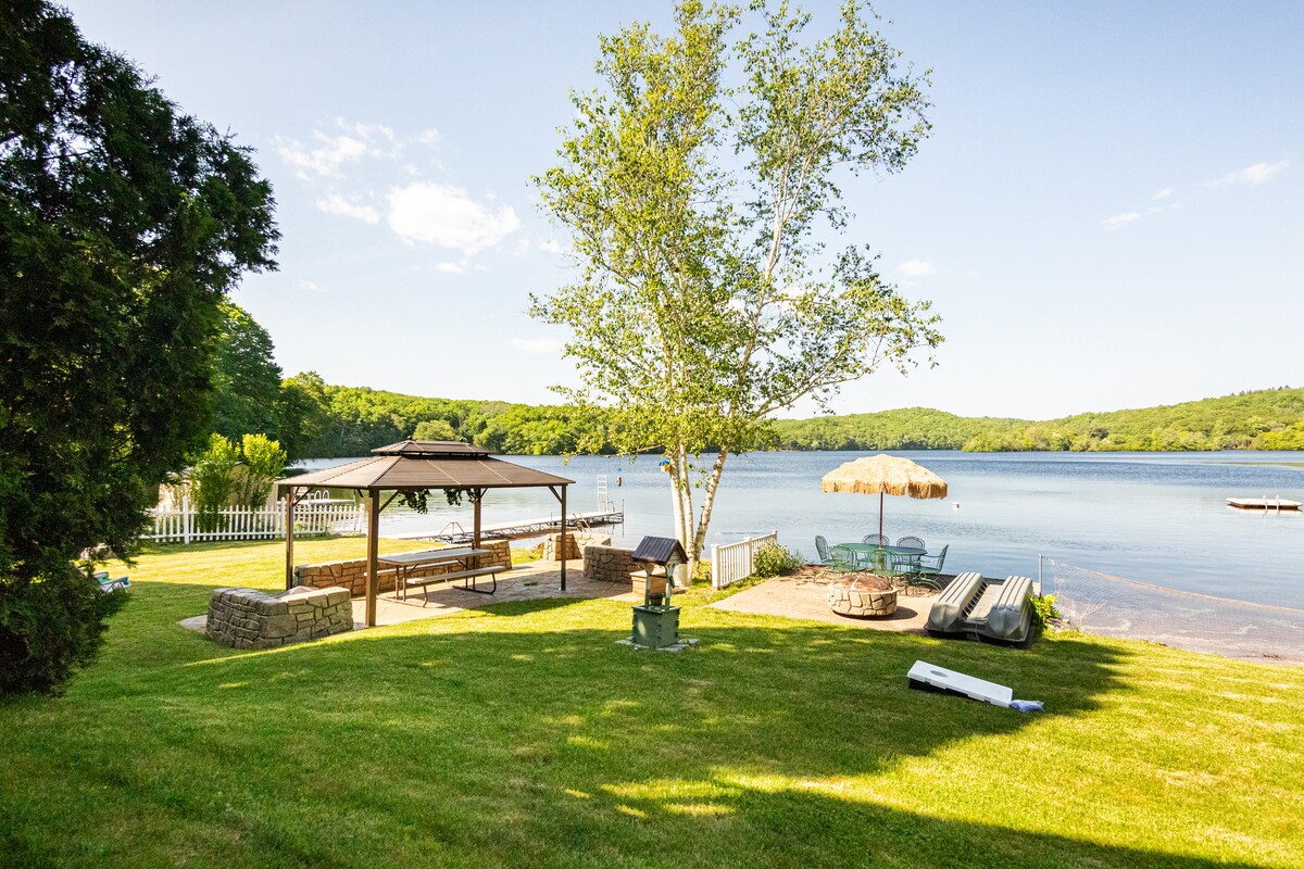 A spacious outdoor area is highlighted by a gazebo and a thatched umbrella resting on a grassy lawn. Nearby, lounge chairs and a stone fire pit are positioned near the serene lake, which is surrounded by lush green hills under a clear blue sky.
