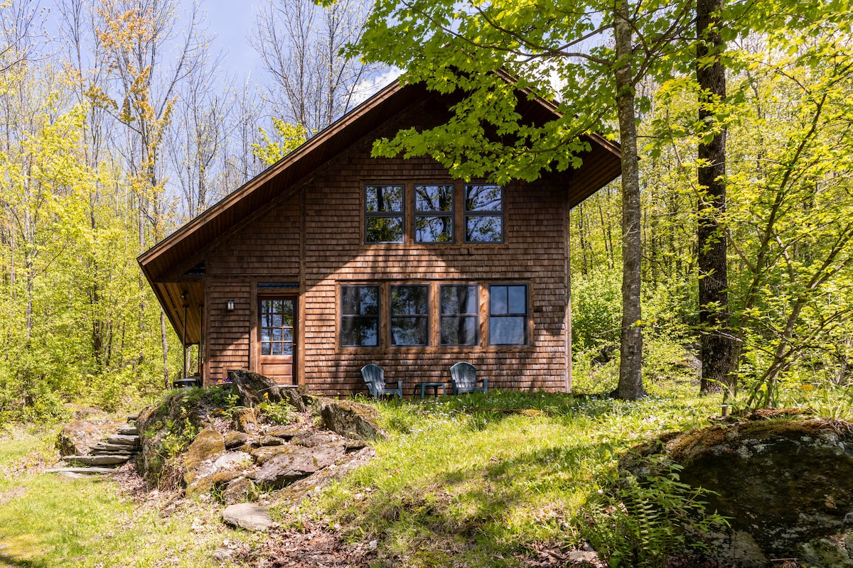 A rustic cabin with a wooden exterior is surrounded by lush greenery. Large windows allow ample natural light, and two patio chairs are positioned on a stone pathway leading to the entrance. The natural landscape enhances the serene setting.
