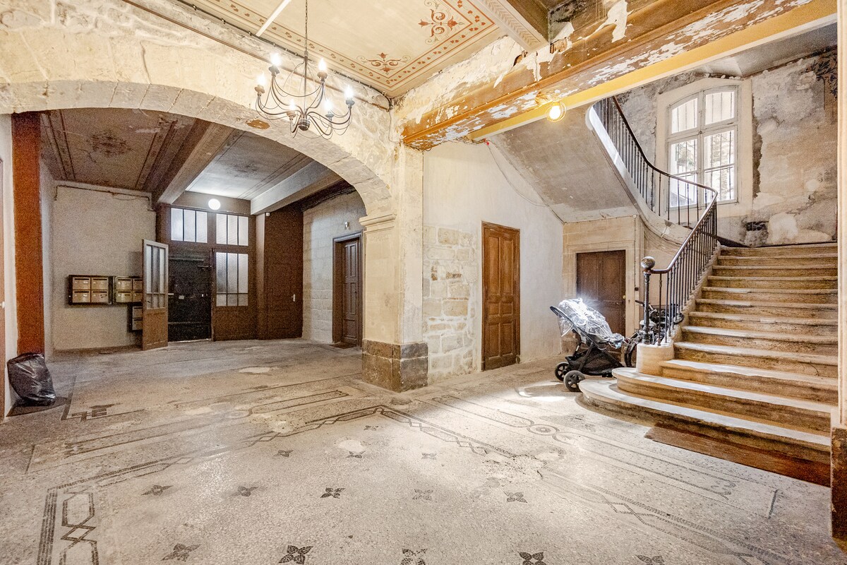 The entryway features high ceilings with decorative elements and a large staircase leading to the upper level. Natural light enters through tall windows, illuminating the stone walls and intricate floor patterns. An arched doorway and wooden doors create an inviting transition to the main living areas.