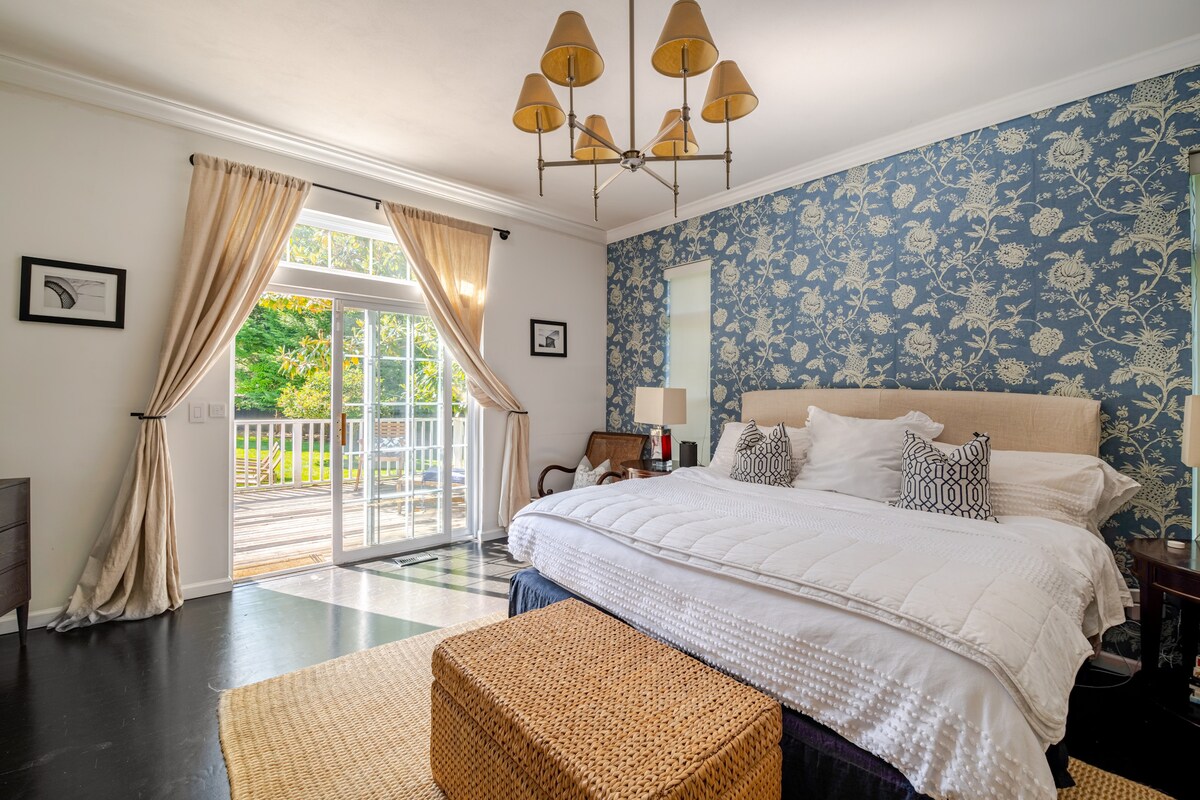 A spacious bedroom is highlighted by a large bed dressed in crisp white linens. A decorative chest serves as a bedside table. Two sets of glass doors lead to an outdoor deck, framed by flowing curtains and accented by a floral wallpaper.