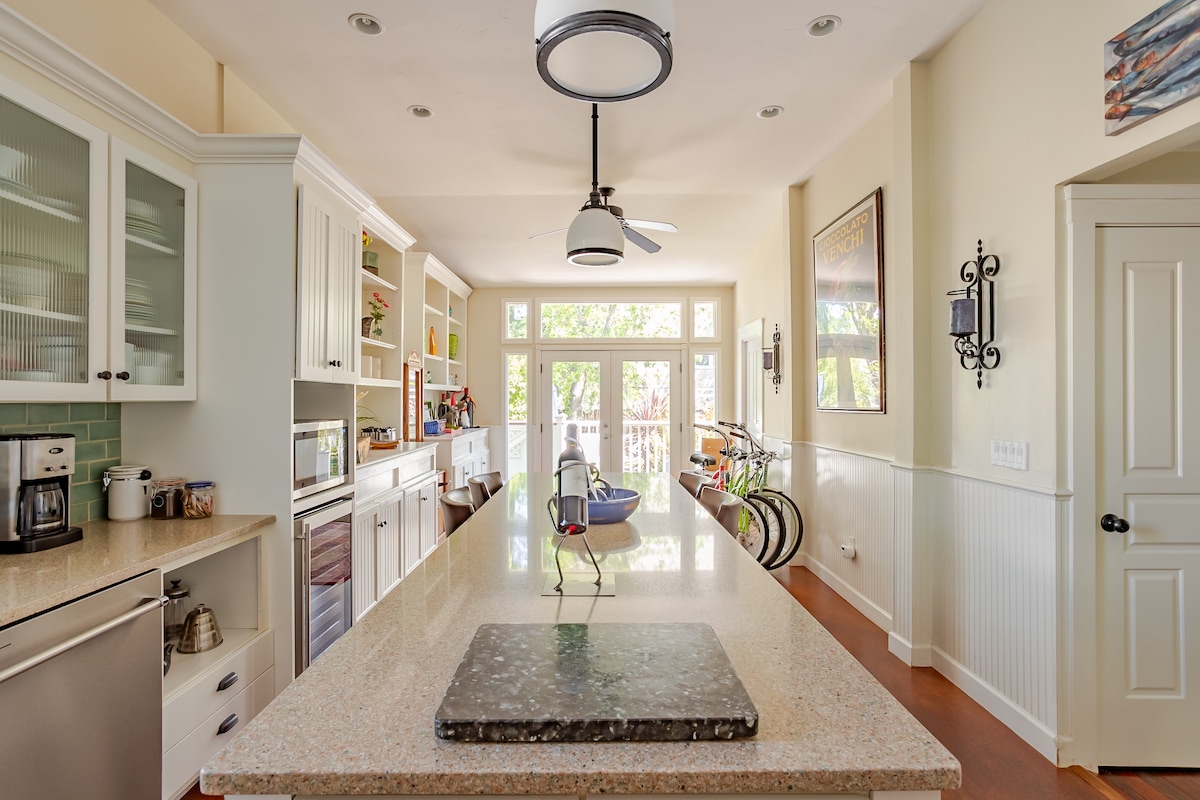 A spacious kitchen features a long central island with a stone countertop, illuminated by a modern light fixture. Natural light fills the room through large windows, highlighting neatly organized cabinets and appliances, while a door leads to an outdoor area. Bicycles are visible, adding a hint of outdoor adventure.