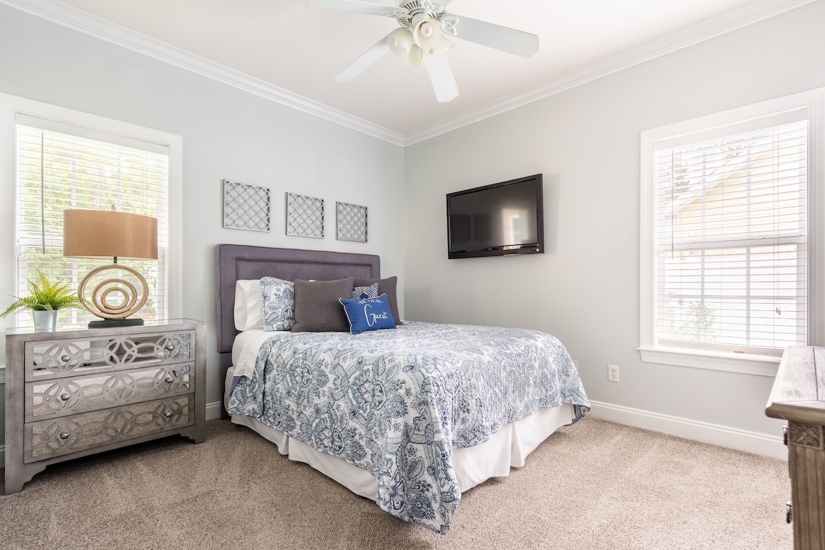 A well-appointed bedroom features a plush bed dressed in blue and white bedding. Two windows provide natural light, while a television is mounted on the wall. A decorative lamp and a stylish dresser complete the space, adding character to the room.