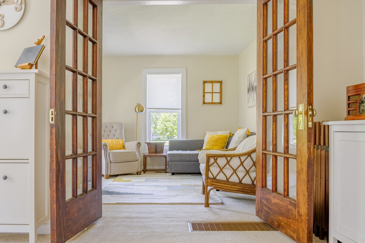 The living room features a cozy seating arrangement, including a gray sofa and an armchair set against a light wall. Natural light is invited through the large windows, with a soft area rug anchoring the space. Decorative elements enhance the welcoming ambiance.