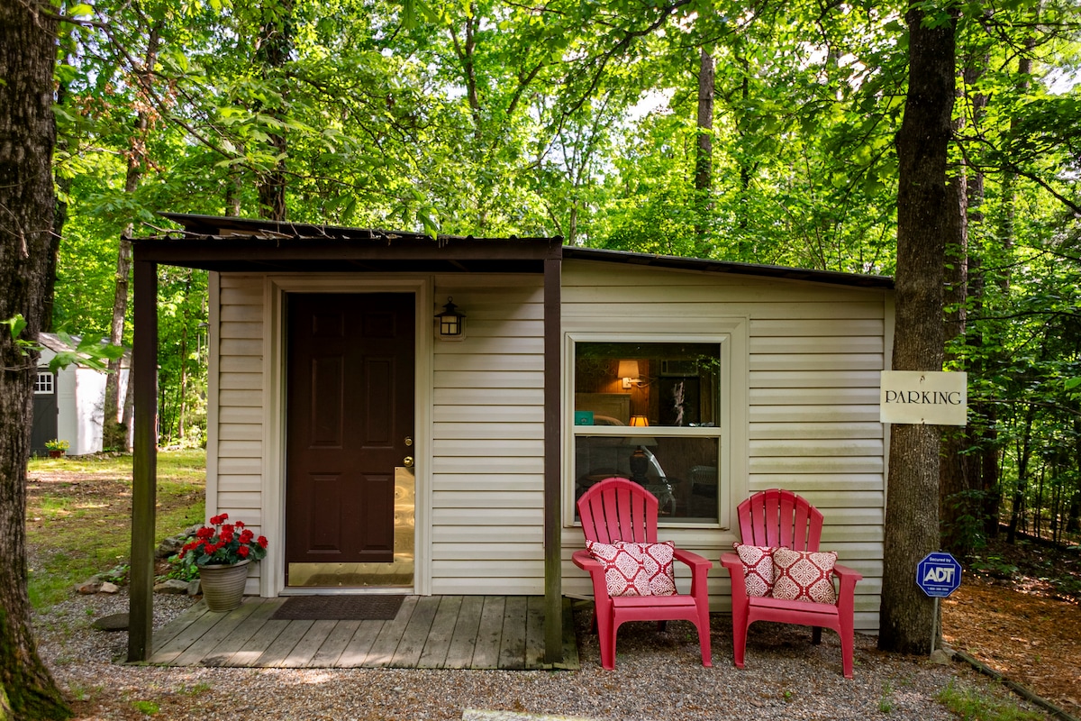 A charming cottage entrance is framed by lush greenery, featuring a welcoming porch with two pink chairs adorned with decorative cushions. A sign indicates designated parking, and a door with a lantern light leads inside, inviting guests to explore the tranquil woodland surroundings.