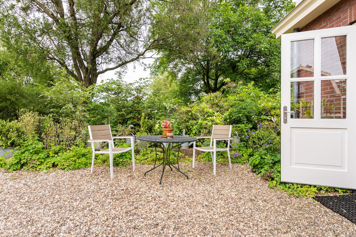 An outdoor seating area features a small round table with a decorative centerpiece, surrounded by two chairs. Lush greenery and flowering plants provide a natural backdrop, enhancing the outdoor ambiance. The entrance door is partially open, leading into the apartment.